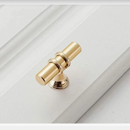 A small metal furniture or cabinet handle with a modern and elegant T-shaped design, featuring a bright gold color and carefully polished finish, mounted on a white wooden surface with classic engraved edges